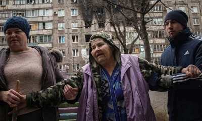 Gente ayudando a una anciana a caminar por la calle junto a un edificio de apartamentos alcanzado por proyectiles en Mariúpol, Ucrania. 